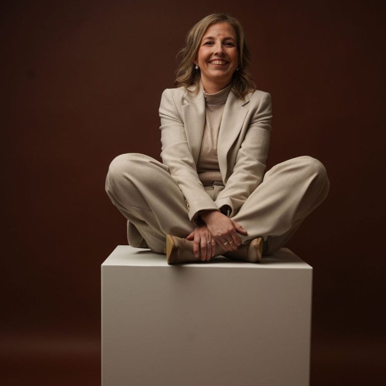 Vrouw in een lichte broek en blazer, zittend op een witte sokkel met een glimlach.